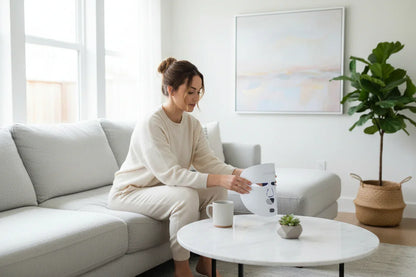Woman in cozy outfit sitting on gray sofa holding LED therapy face mask in bright modern living room
